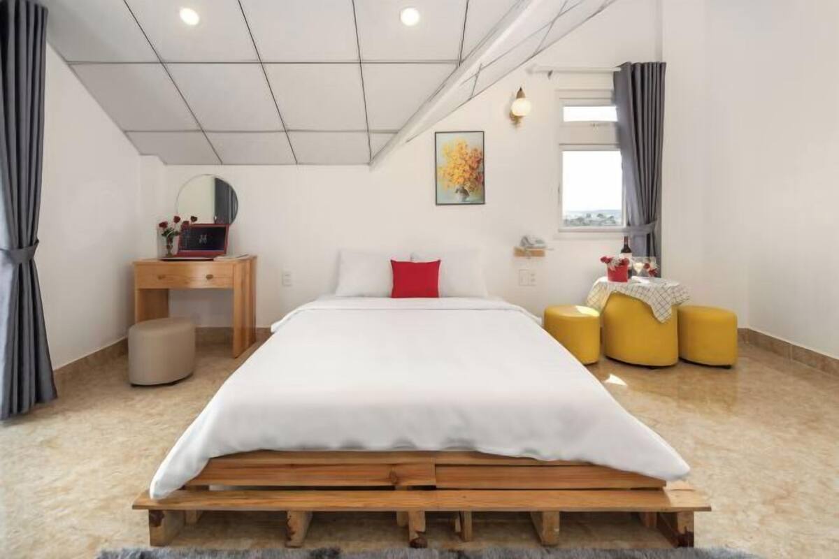 Une chambre à coucher avec un lit en bois, une table de chevet, un miroir, un oreiller rouge et une fenêtre avec des rideaux.