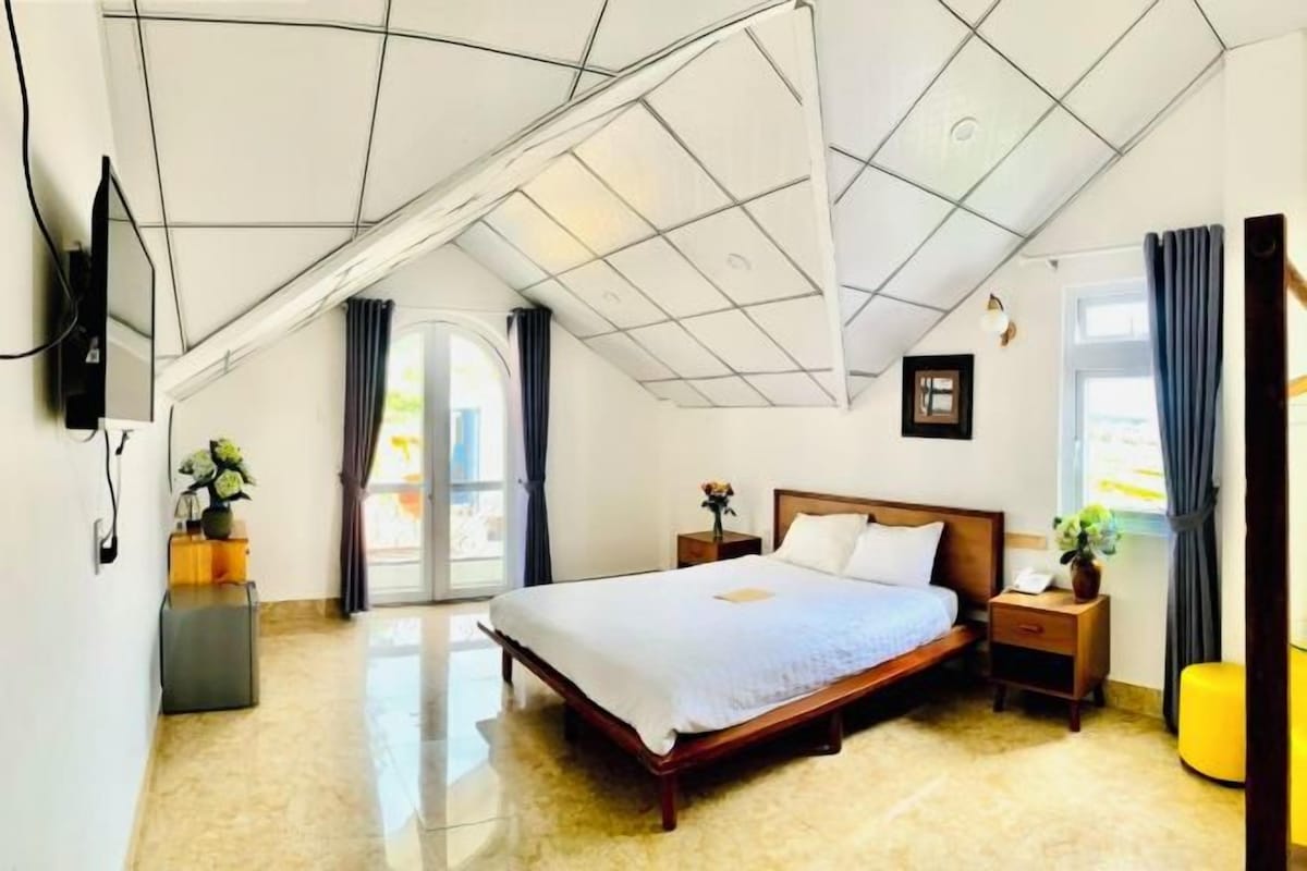 A modern bedroom with a large bed, a flat-screen TV mounted on the wall, and a window with curtains.