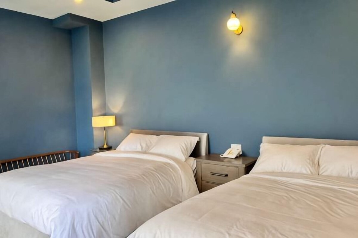 A hotel room with two beds, a nightstand with a lamp, and a wall-mounted light fixture.