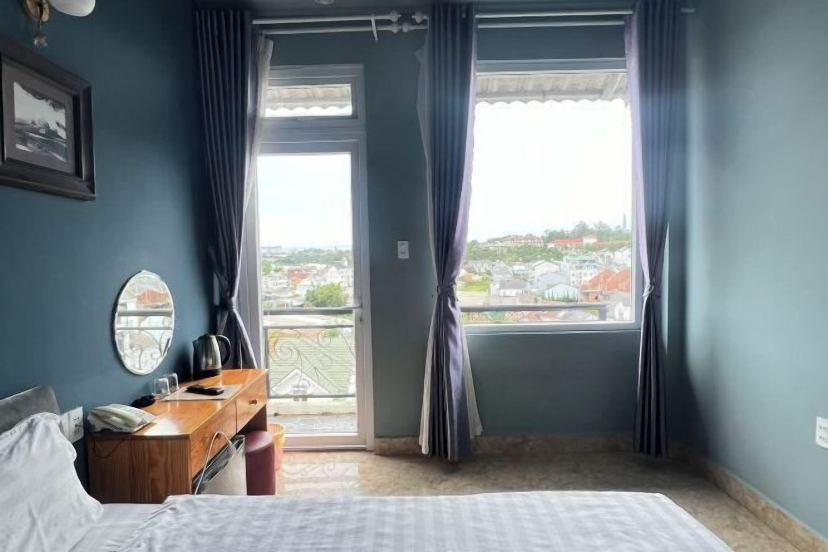 Une chambre à coucher comprenant un lit, un bureau, une chaise, une fenêtre avec des rideaux et une vue sur les bâtiments environnants.