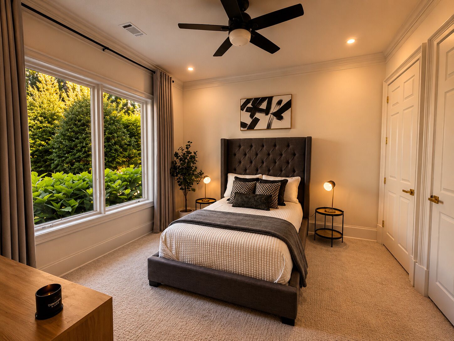 A bedroom with a large window, a ceiling fan, a bed with a dark headboard, and a bedside table with a candle.