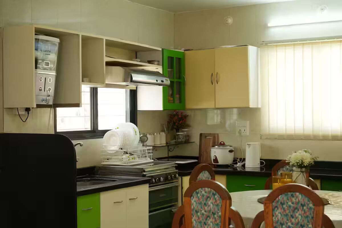 Uma cozinha compacta com fogão, pia e armários verdes. Há uma mesa de jantar com cadeiras e um vaso de flores.