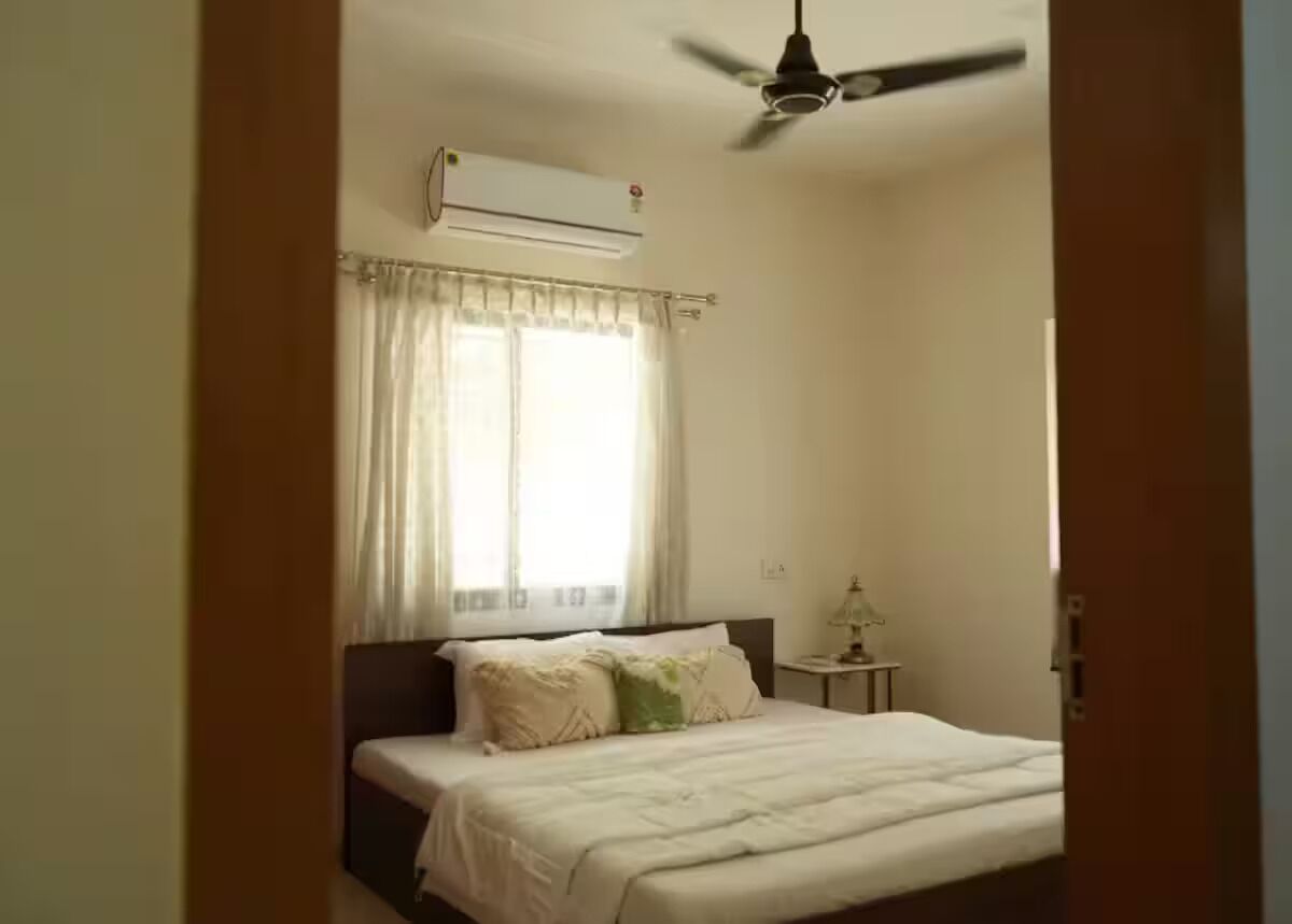 Quarto com cama, ventilador de teto, ar condicionado e janela com cortinas.