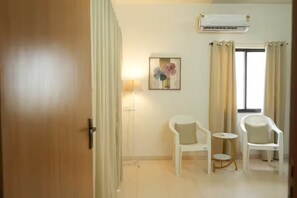 A room with a wooden door, a white air conditioning unit, a small table, two white chairs, and a window with curtains. - Vayavia White House / Where Comfort Feel Like Home (Nashik)