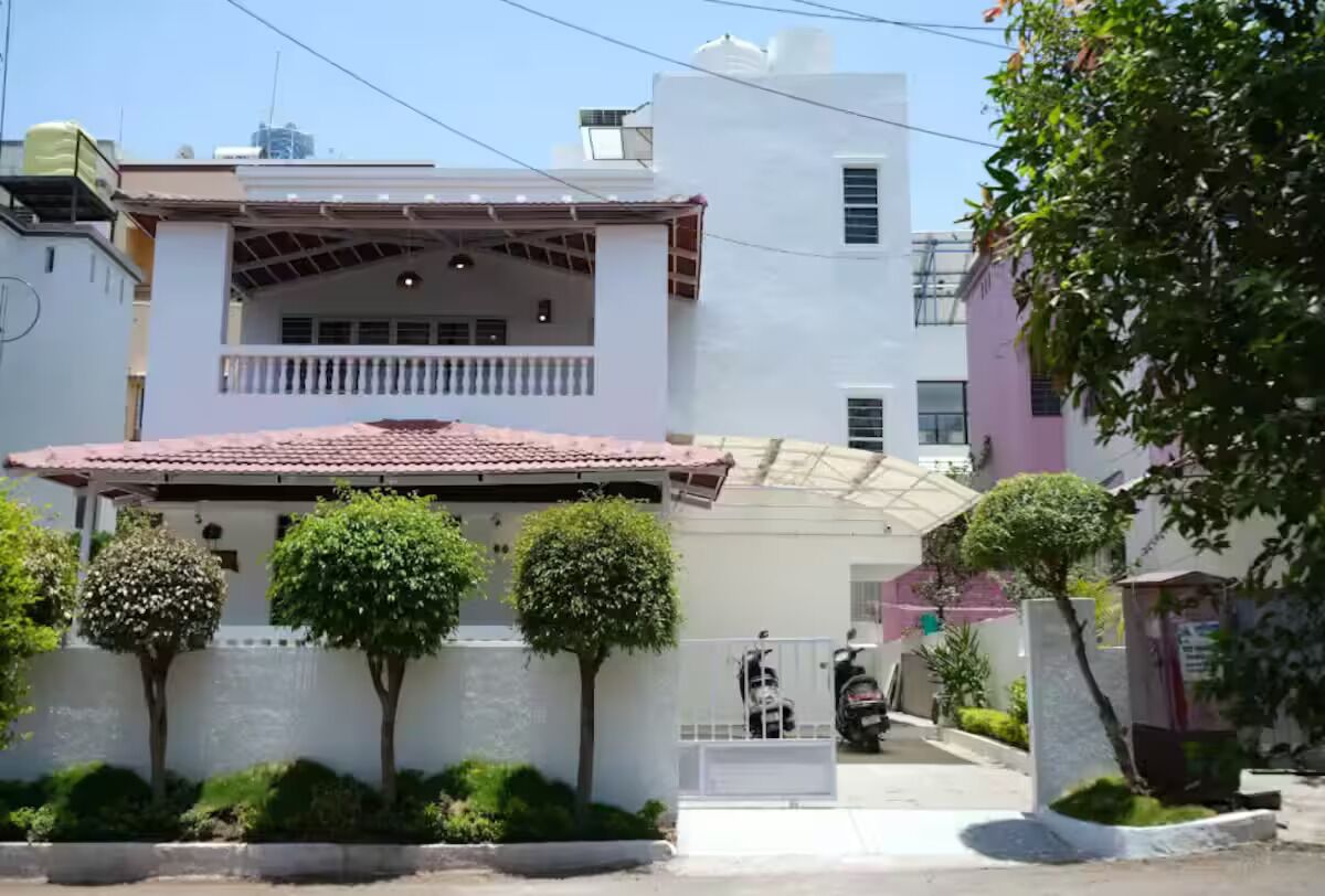 Um edifício residencial com varanda coberta, portão fechado e um pequeno jardim com arbustos aparados.