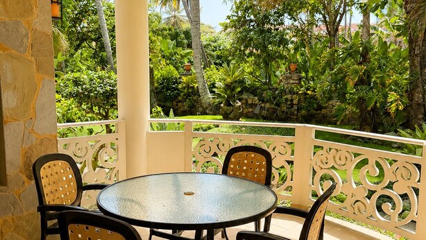 Un área de estar al aire libre con techo, una mesa redonda de vidrio y cuatro sillas, rodeada por una barandilla decorativa y abundante vegetación.