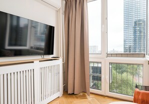 A modern living room with a flat-screen TV mounted on a white wall, a large window with a view of a cityscape, and a wooden floor. - Rondo ONZ Cozy Stay Apartment (Warszawa)