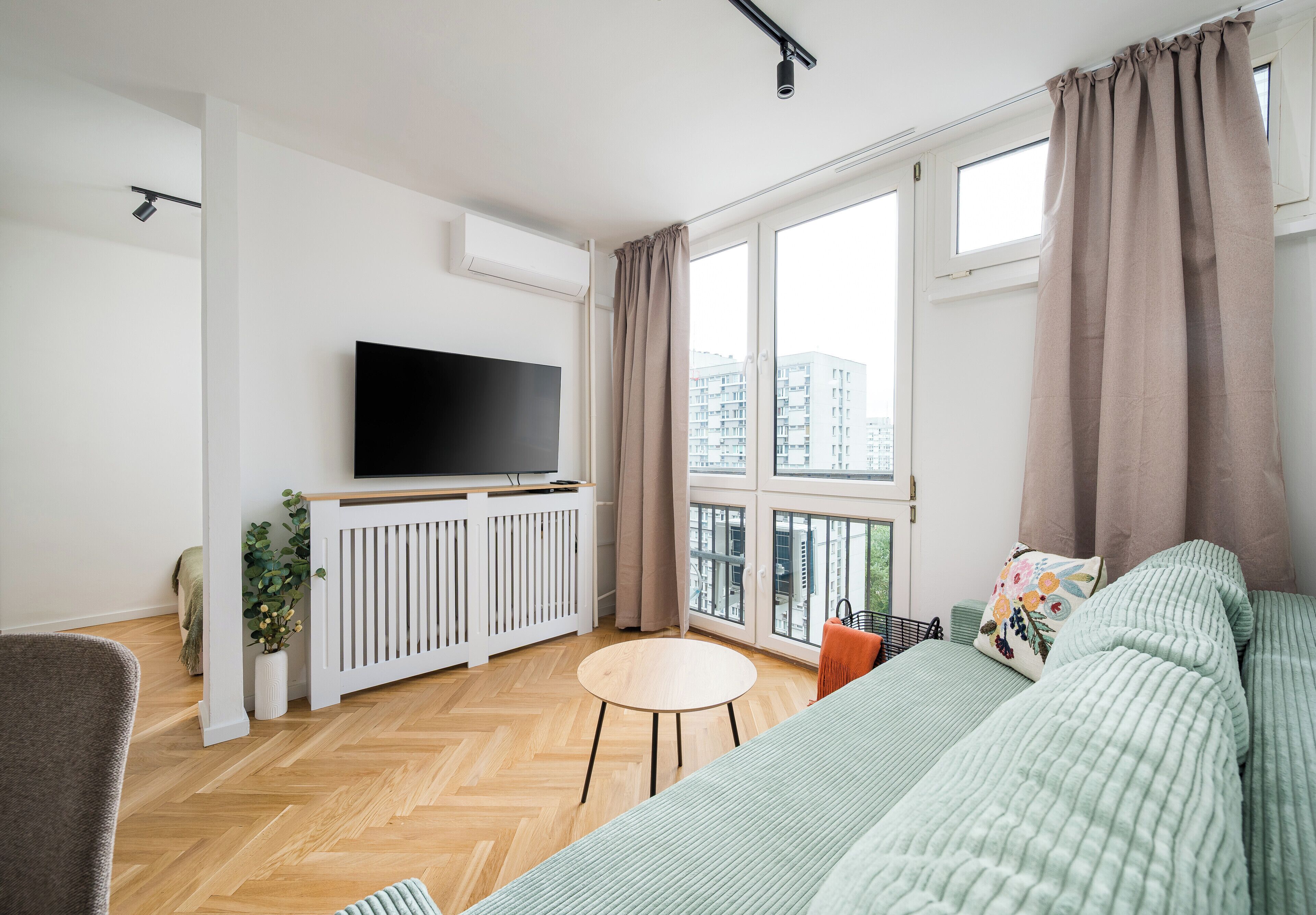 A modern living room with a flat-screen TV, a sofa, a small round table, and large windows with curtains.