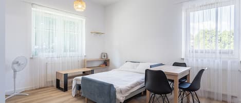 A minimalist bedroom with a bed, bedside table, chair, and a standing fan.
