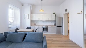 Una cocina moderna con armarios blancos, horno empotrado y refrigerador. En primer plano, una mesa de comedor con sillas y un sofá gris.