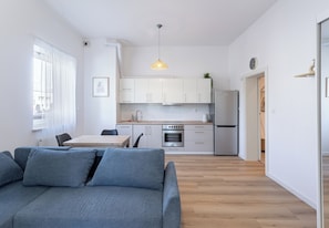 Una cocina moderna con armarios blancos, horno empotrado y refrigerador. En primer plano, una mesa de comedor con sillas y un sofá gris.