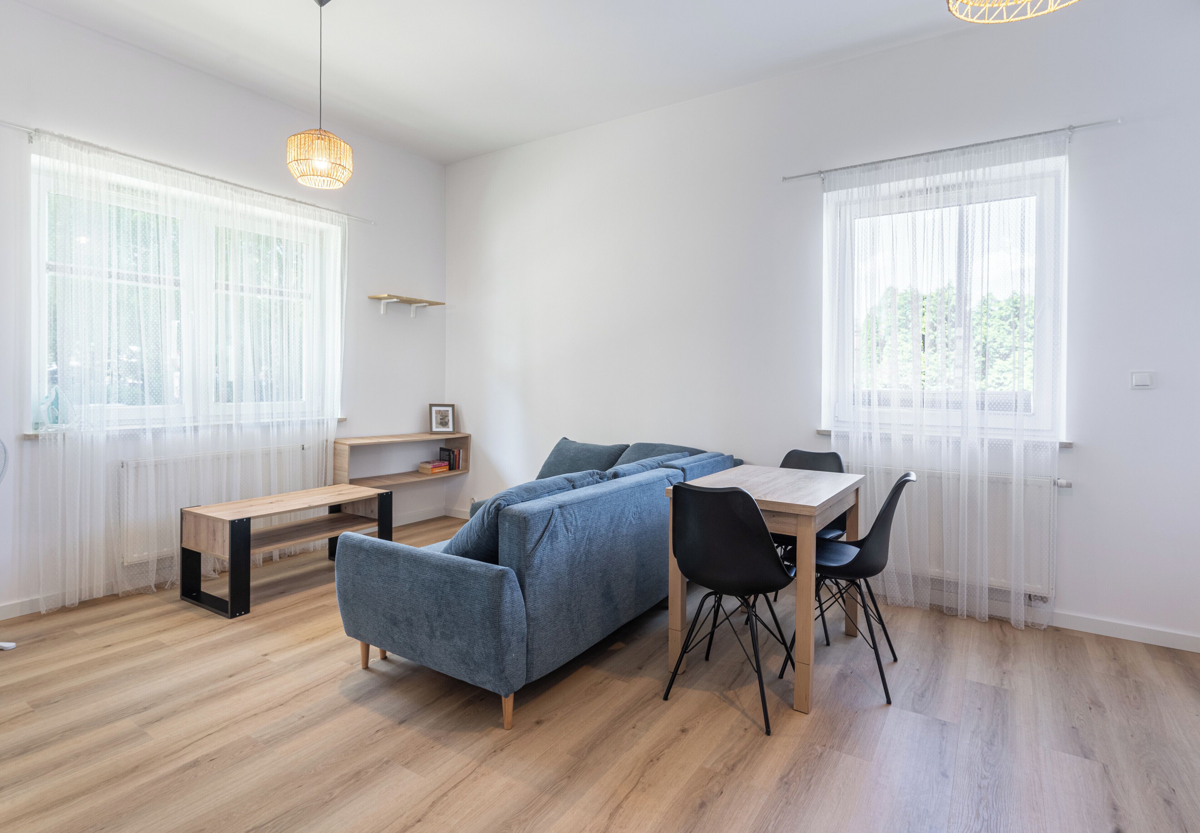 Un salon moderne avec un canapé bleu, une table à manger en bois et des chaises noires. Il y a deux fenêtres avec des rideaux légers, un luminaire suspendu et une petite étagère avec des livres.