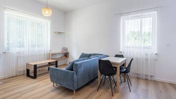 Un salon moderne avec un canapé bleu, une table à manger en bois et des chaises noires. Il y a deux fenêtres avec des rideaux légers, un luminaire suspendu et une petite étagère avec des livres.
