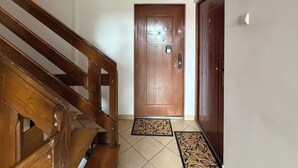 Una escalera de madera que conduce a una puerta con un cartel con número, un felpudo y un interruptor de luz en la pared.