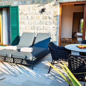 Pared de piedra con un sofá y sillones, una mesa con fruta, y una puerta con contraventanas verdes.