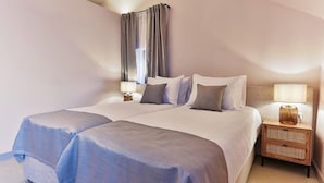 Ein Doppelbett mit heller Bettdecke, ein Nachttisch mit Lampe und ein Fenster mit Vorhängen.
