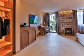 A hotel room with a wooden wardrobe, a television, a small table with a chair, a balcony with a stone wall, and a sofa. - Boutique Hotel Ruzmarin SPA & Garden (Budva)