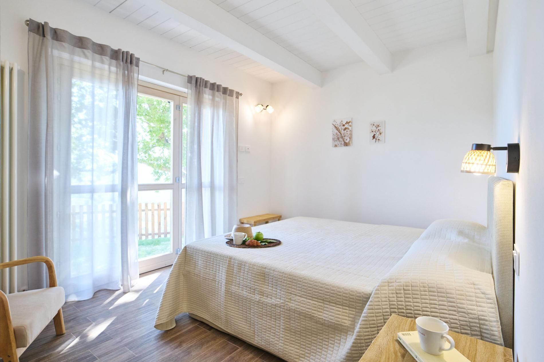 A hotel room with a bed, a chair, a table with a cup and a book, and a window with curtains.