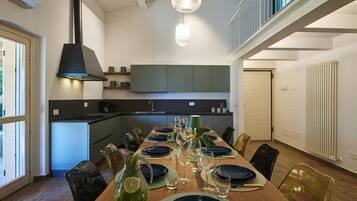 A modern dining area with a long table set for a meal, a kitchen in the background, and a staircase leading to an upper level.