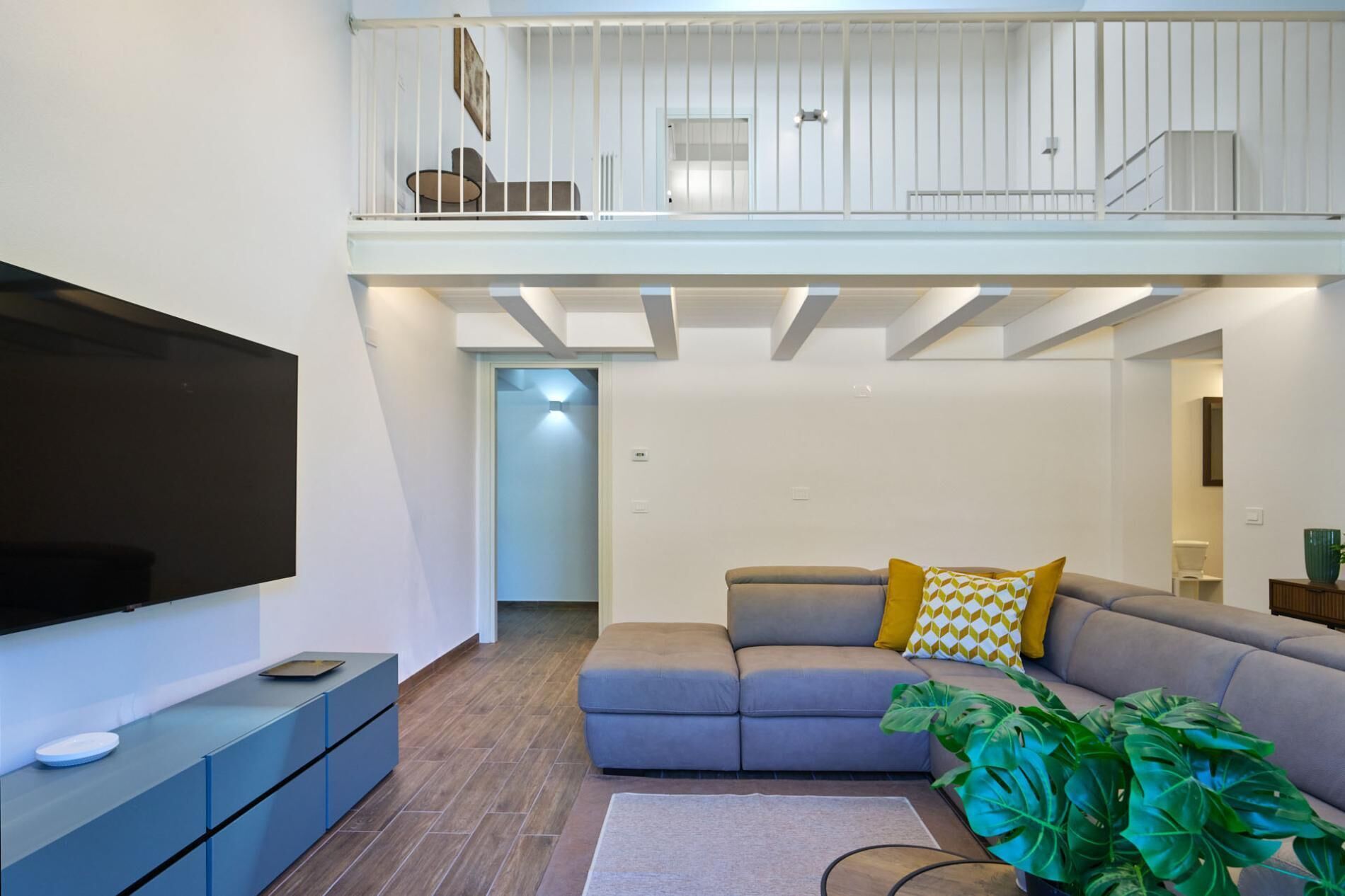 A modern living room with a flat-screen TV, a grey sofa with patterned cushions, a coffee table, and a large green plant.