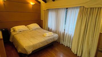 Una habitación de hotel con una cama, suelo de madera, una mesita de noche y un ventanal con cortinas.