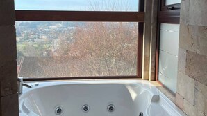 A bathroom with a large bathtub, a window with a view of a cityscape, and stone walls. - Hotel Peregrino Suites (Concepción)