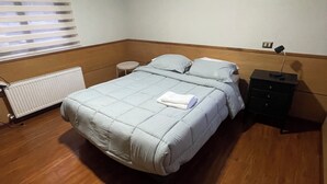 A bedroom with a bed, a nightstand, a radiator, a window with blinds, and a wall-mounted light fixture. - Hotel Peregrino Suites (Concepción)
