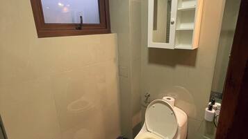Un baño con inodoro, un bote de basura pequeño, una ventana y un espejo.