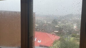 A window with a view of a red-roofed building and a green area, with raindrops on the glass. - Hotel Peregrino Suites (Concepción)