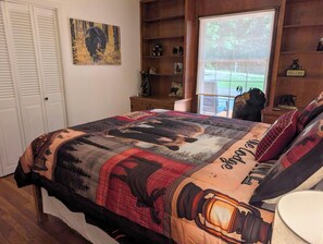 A bedroom with a large bed, a window with a view of the outdoors, a wooden headboard, and a wall with a bear-themed artwork.