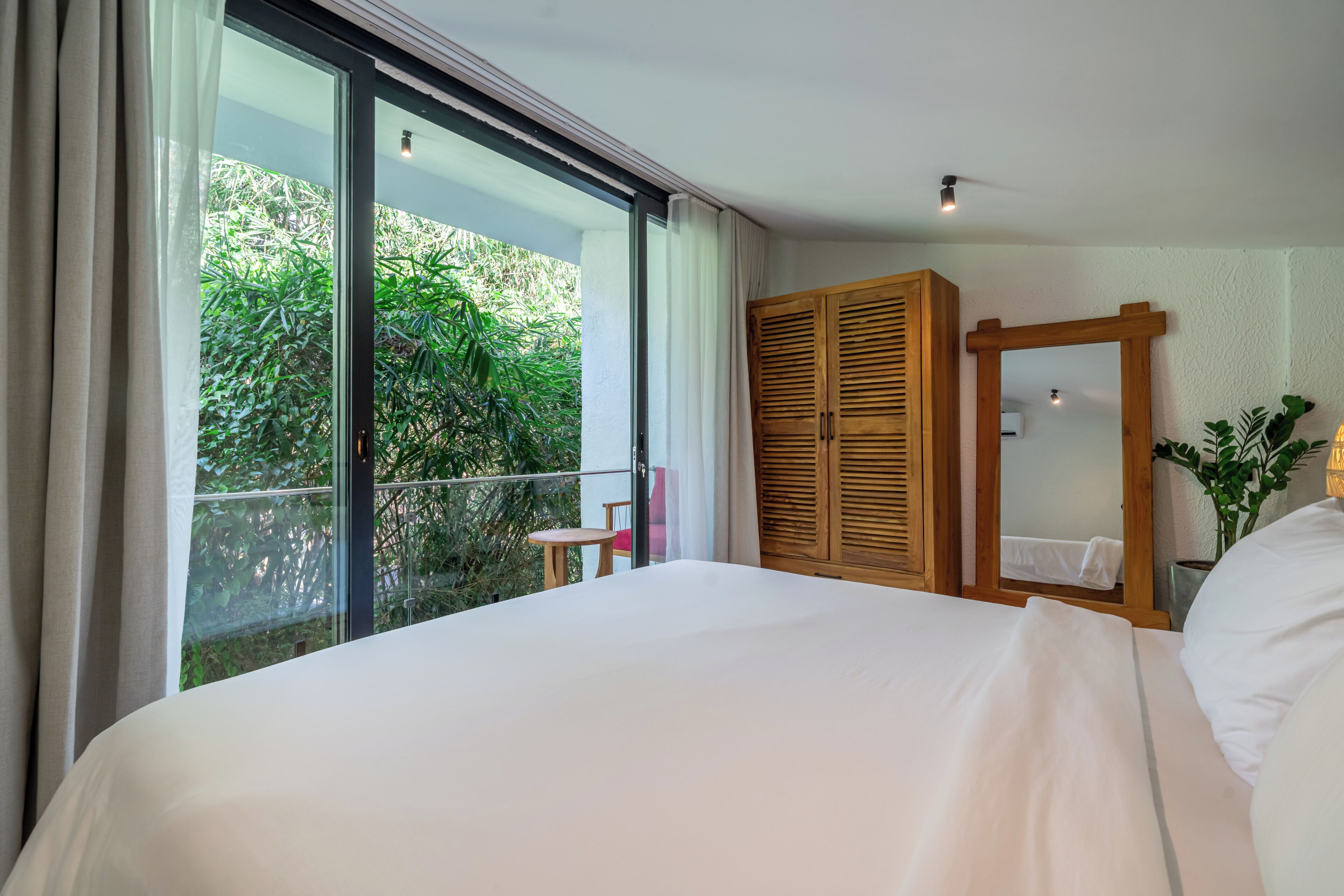 Un dormitorio con una cama grande, un armario de madera, una mesita pequeña y un balcón con vistas a la vegetación.