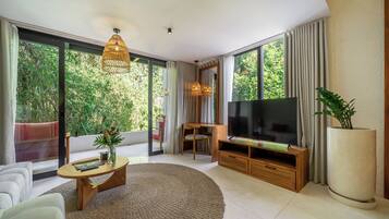 A modern living room with a large TV, a coffee table, and a sofa. There is a large window with curtains, a plant, and a view of greenery outside.