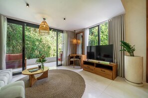 A modern living room with a large TV, a coffee table, and a sofa. There is a large window with curtains, a plant, and a view of greenery outside. - Sacred Jungle Villas & Apartments (Gianyar)