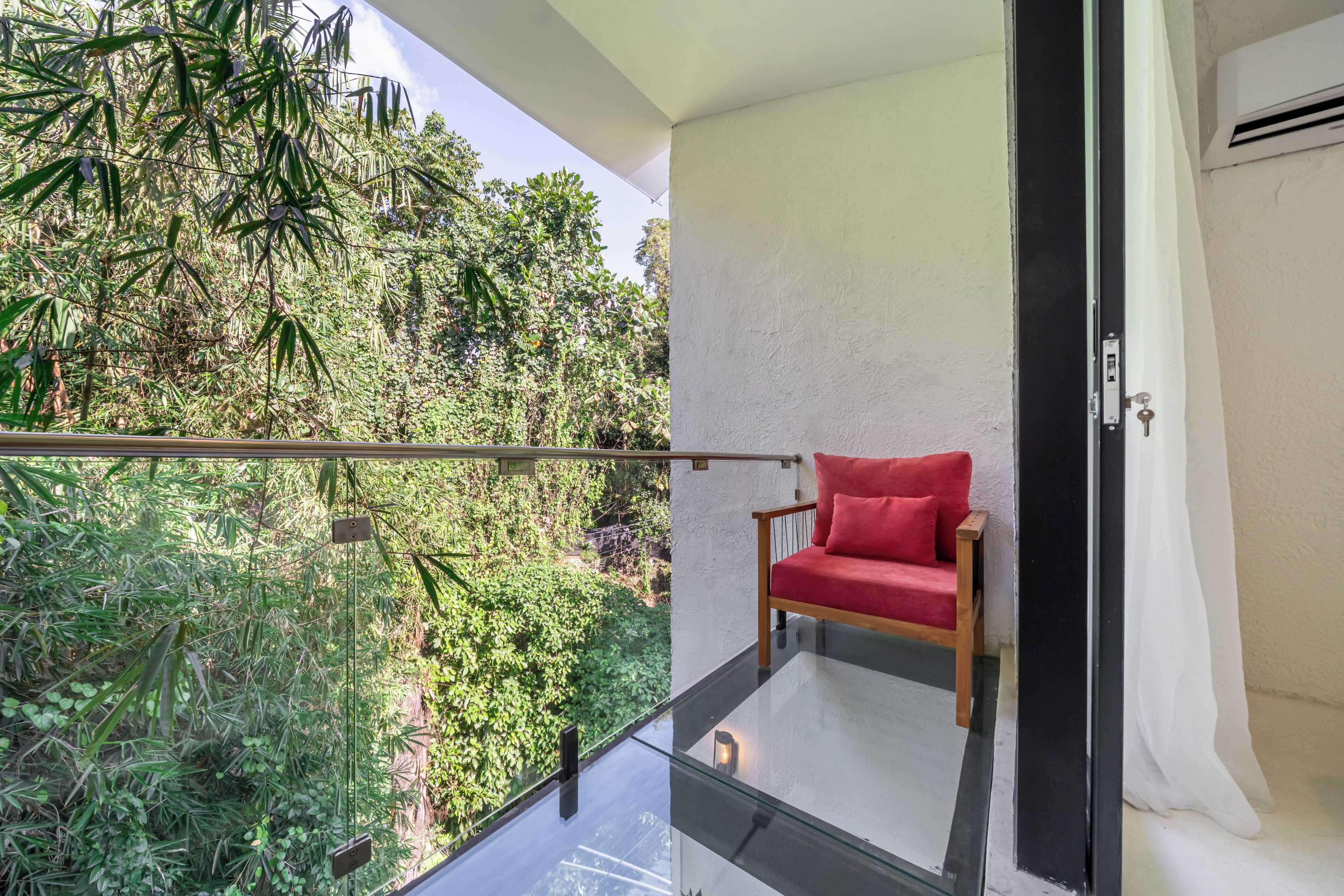 A balcony with a glass railing overlooking dense foliage, a red cushioned chair, and a white wall with a door.