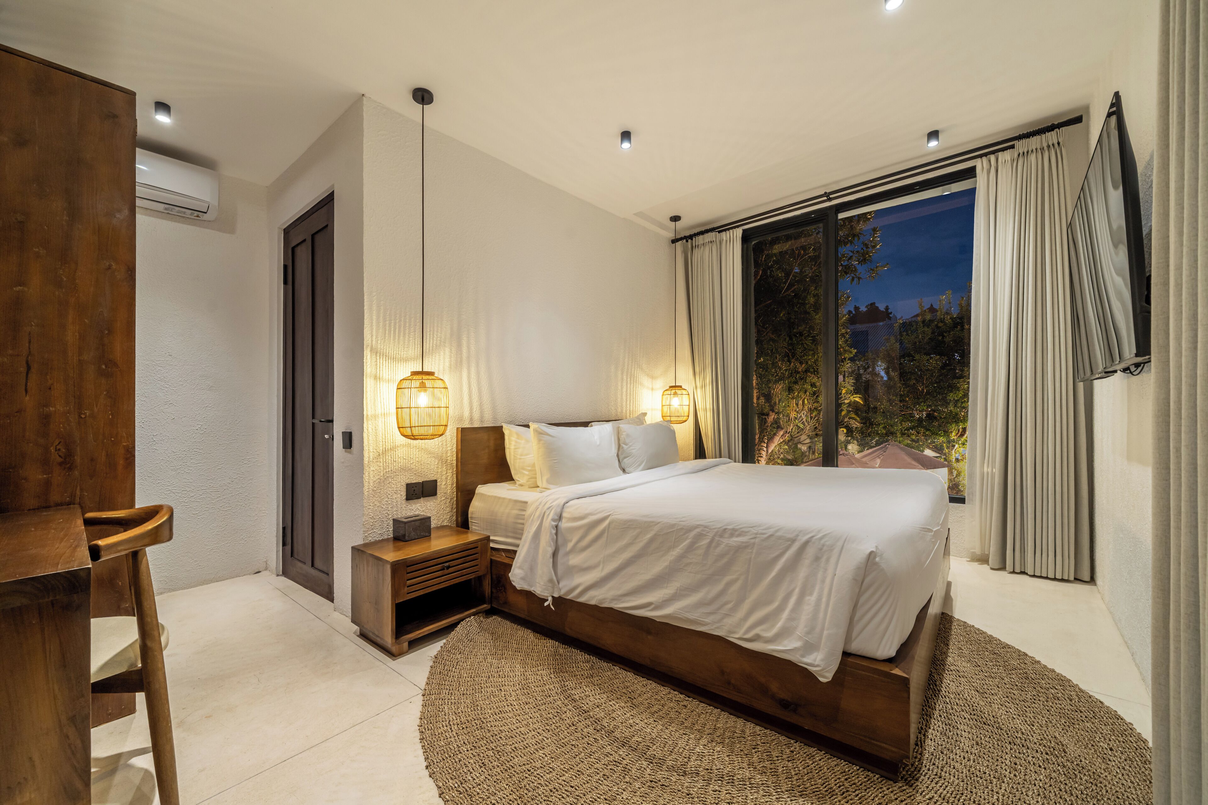 Een moderne slaapkamer met een groot bed, nachtlampjes, een houten bureau en uitzicht op het groen door een groot raam.