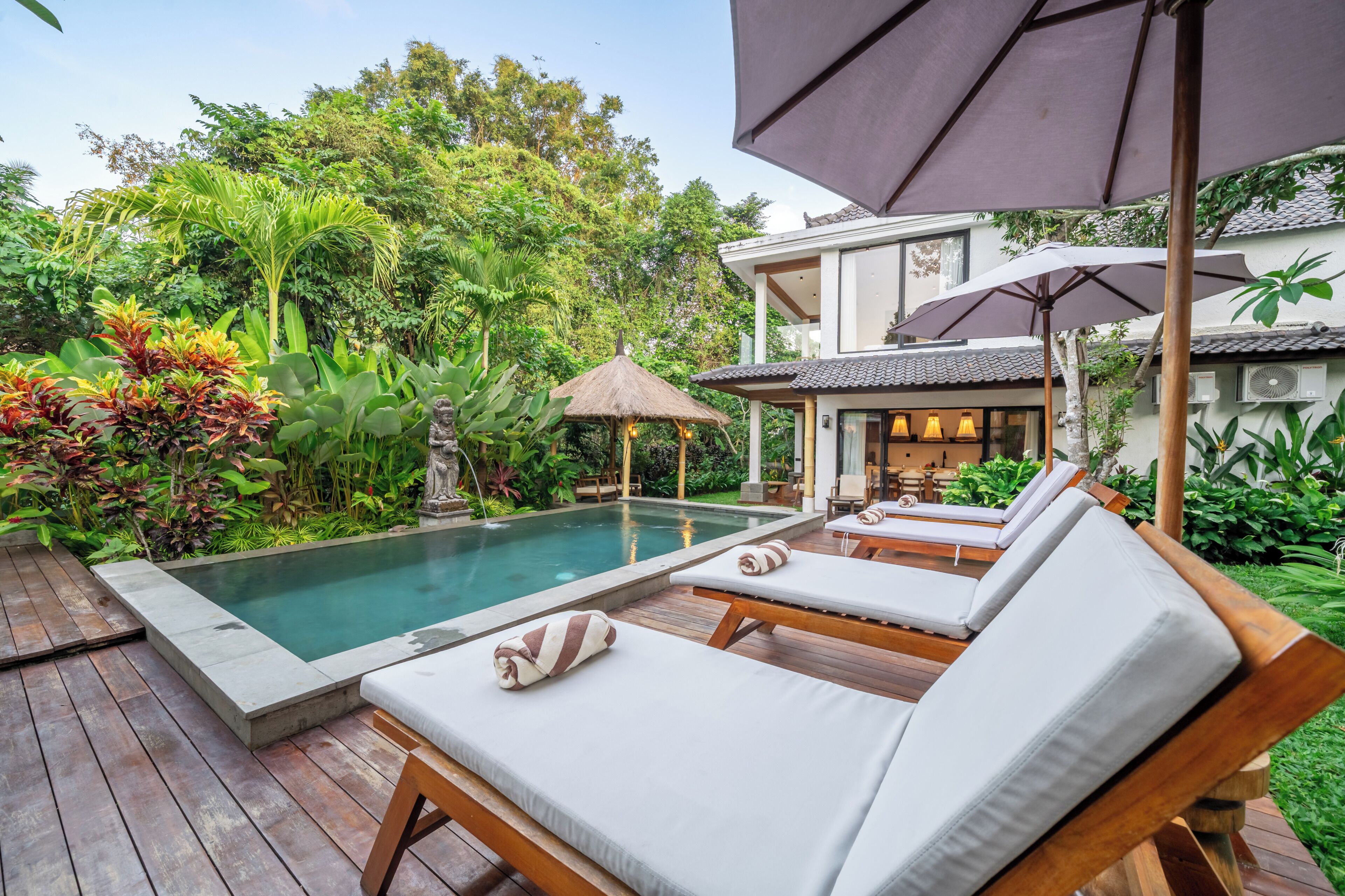 Zona junto a la piscina con sillones de descanso, una sombrilla de paja y exuberante follaje.