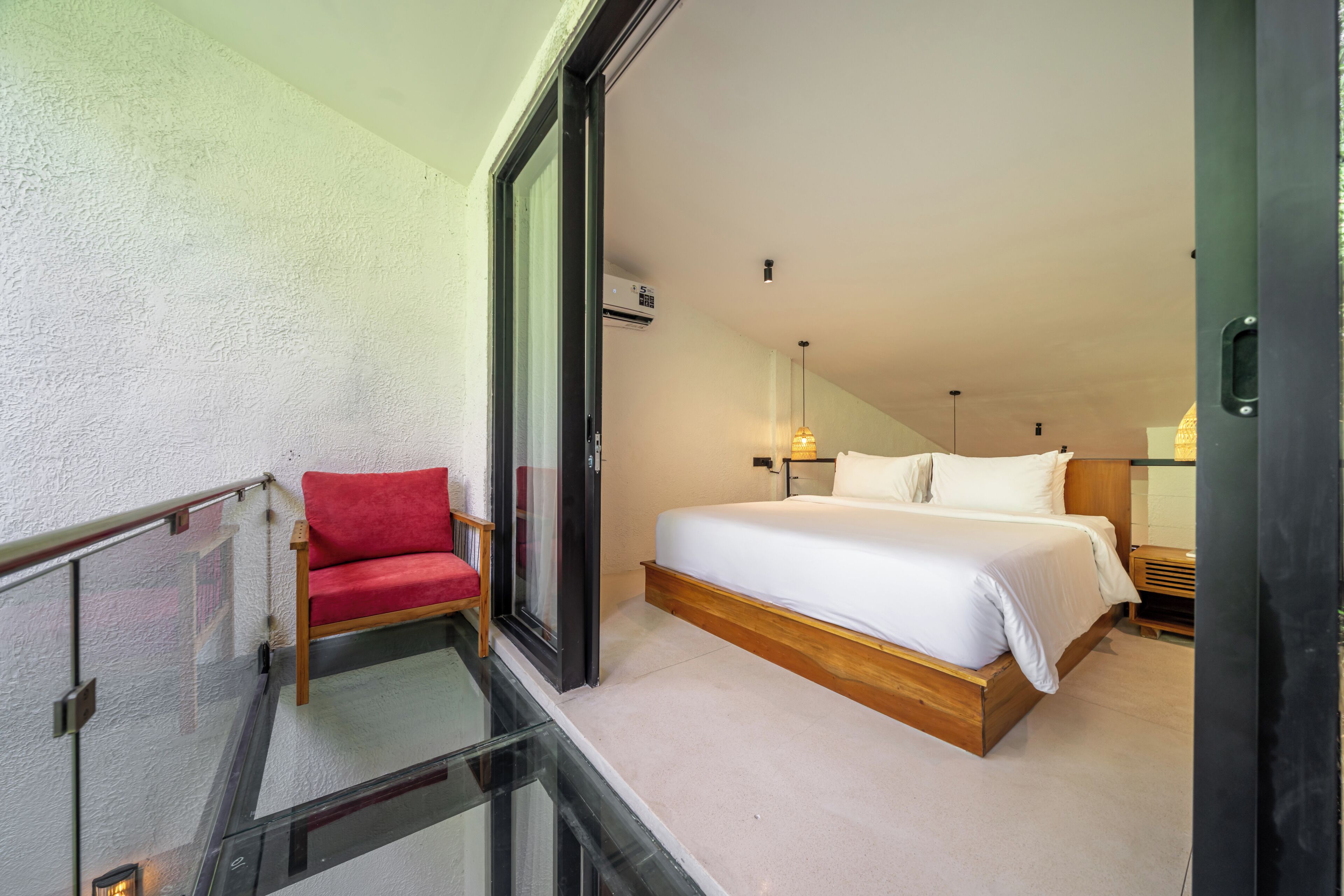A hotel room with a bed, a red chair, a glass balcony, and a ceiling fan.