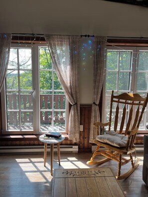A room with a rocking chair, a small table, and a view of a wooden deck through large windows.