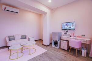 A modern, pink-themed room with a sofa, two chairs, a desk with a computer, a television, and an air conditioning unit.