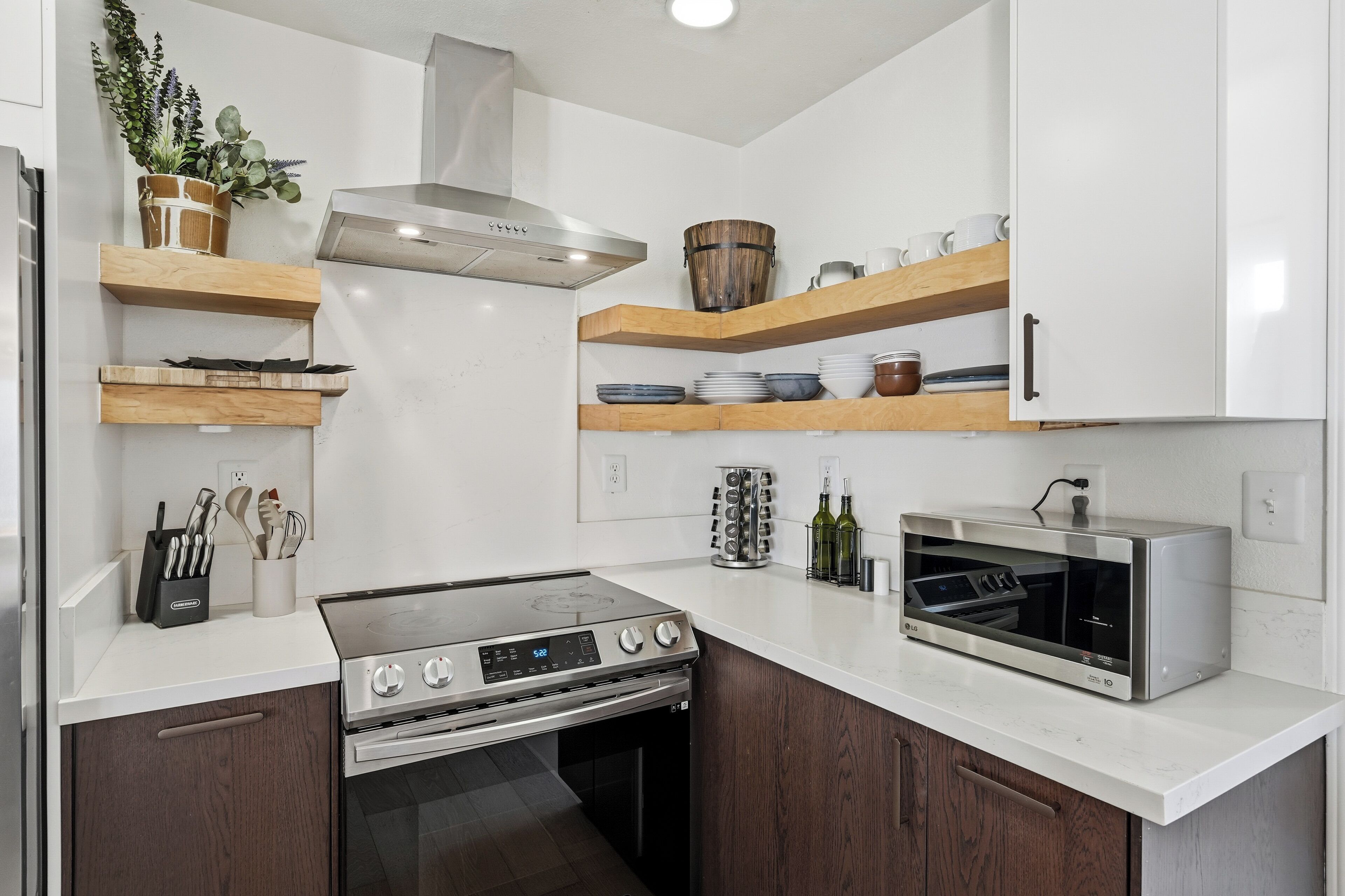 Una cocina moderna con estufa, horno y microondas de acero inoxidable, estantes de madera y una planta en maceta.