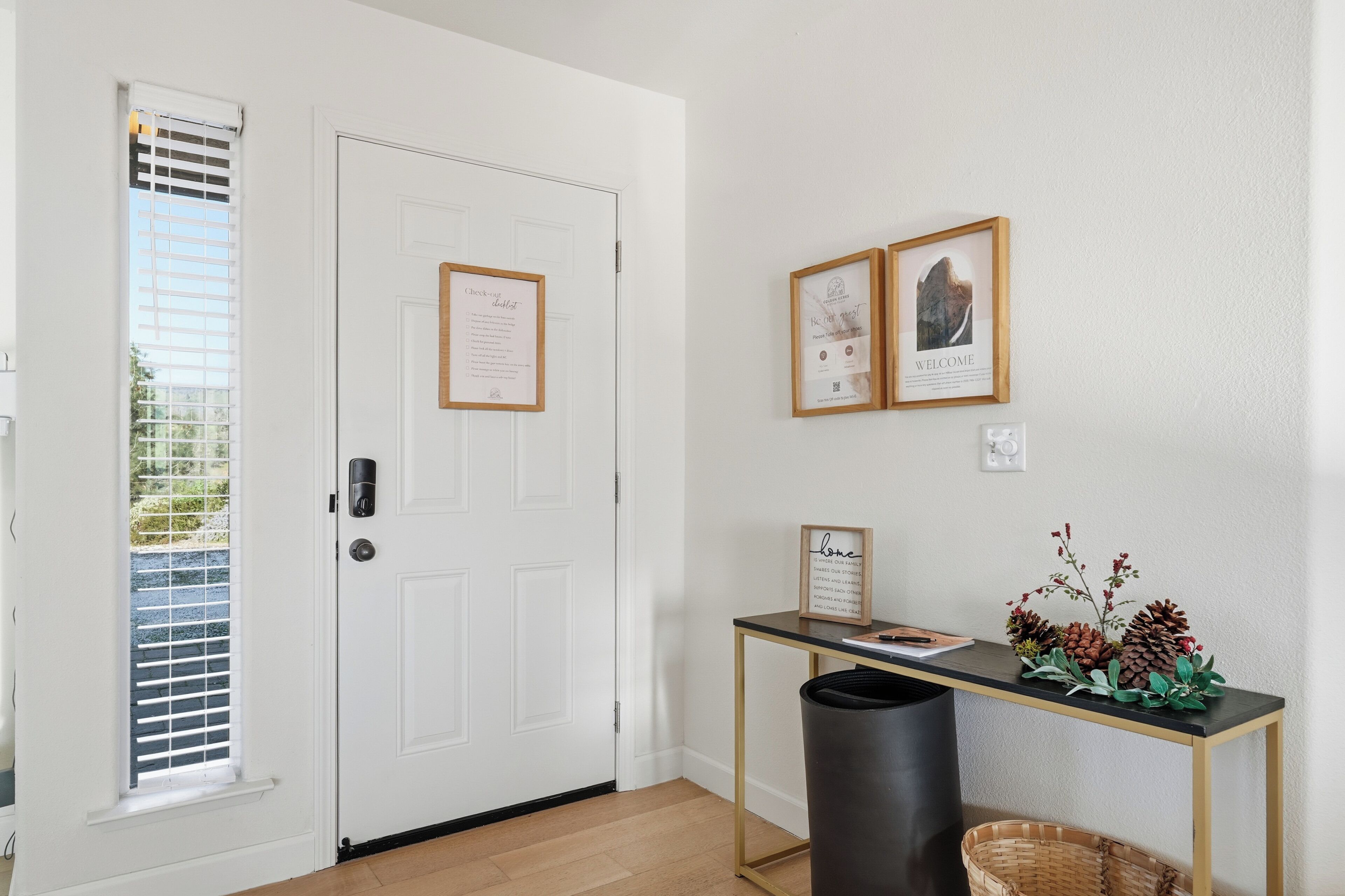 A white door with a notice, a framed welcome sign, a small table with a plant, and a trash bin.