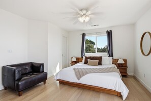 Un dormitorio con una cama, un sofá de cuero oscuro, un ventilador de techo, una ventana con cortinas y piso de madera.