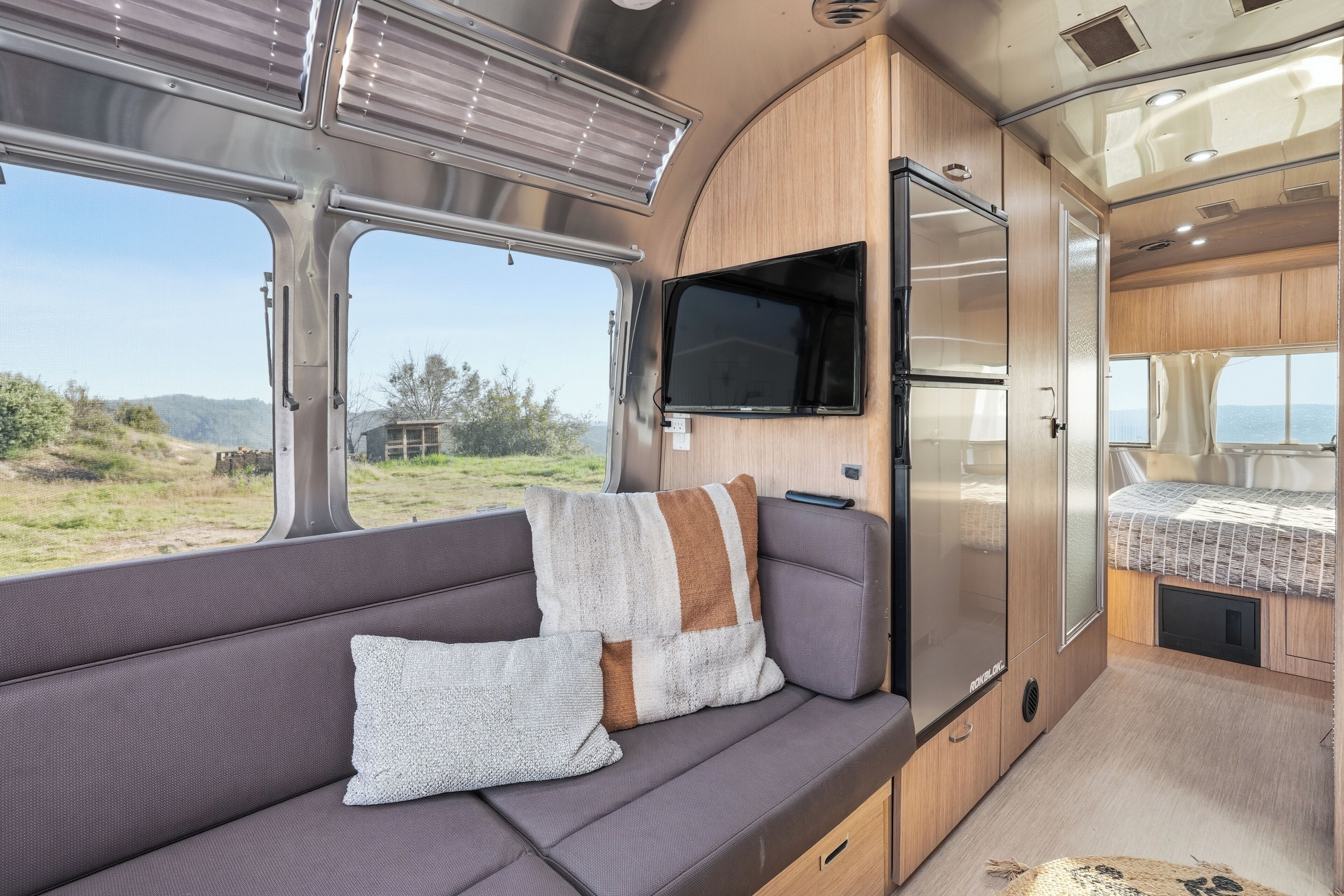 A cozy RV interior with a sofa, a flat-screen TV, a refrigerator, and a window offering a scenic view.