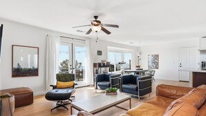 Een moderne woonkamer met een bank, fauteuil en salontafel. Er is een plafondventilator, een eethoek met stoelen en een keukenhoek.