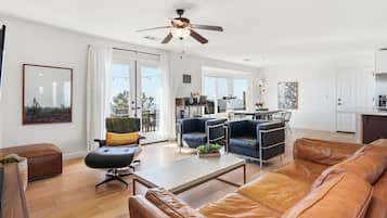A modern living room with a sofa, armchair, and coffee table. There is a ceiling fan, a dining area with chairs, and a kitchen area.