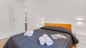 Una cama bien hecha con edredón gris oscuro y dos toallas blancas enrolladas, un cabecero de madera y una revista sobre la cama.