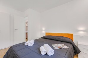 Una cama bien hecha con edredón gris oscuro y dos toallas blancas enrolladas, un cabecero de madera y una revista sobre la cama.
