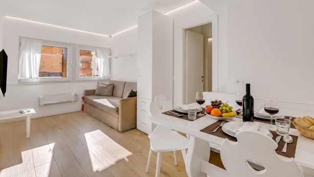 Un área de estar moderna y luminosa, con una mesa de comedor para dos personas, un sofá y vista a la cocina por una puerta abierta.