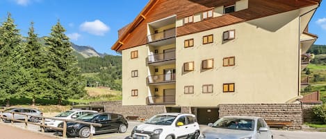 A multi-story hotel with a sloped roof, balconies, and a parking area with several cars.