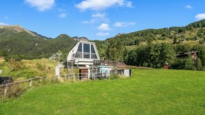 Un refugio de montaña con una amplia estructura de vidrio, rodeado de campos verdes y montañas.